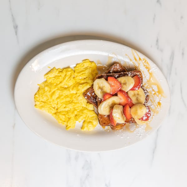 Scrambled eggs and French toast topped with strawberries, bananas, powdered sugar, and syrup.
