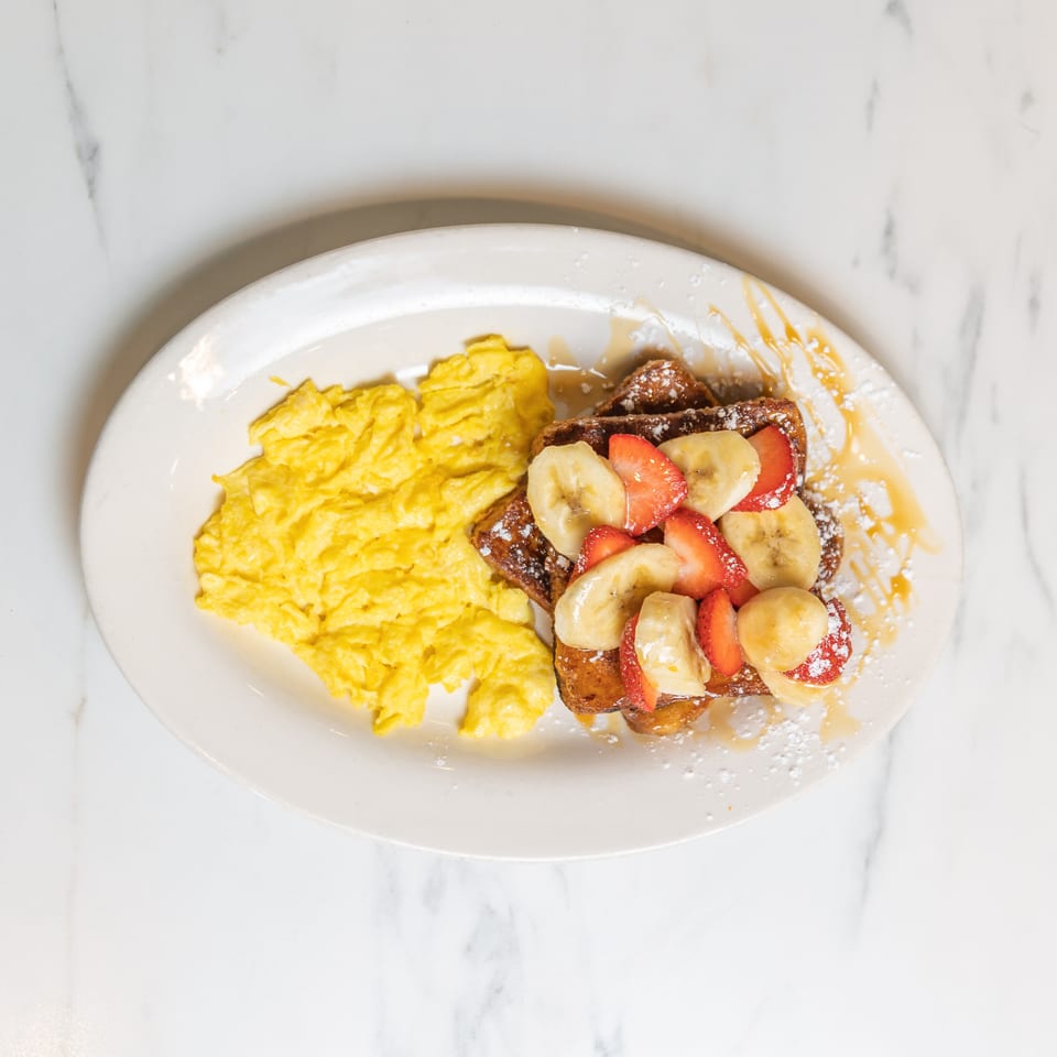 Scrambled eggs and French toast topped with strawberries, bananas, powdered sugar, and syrup.