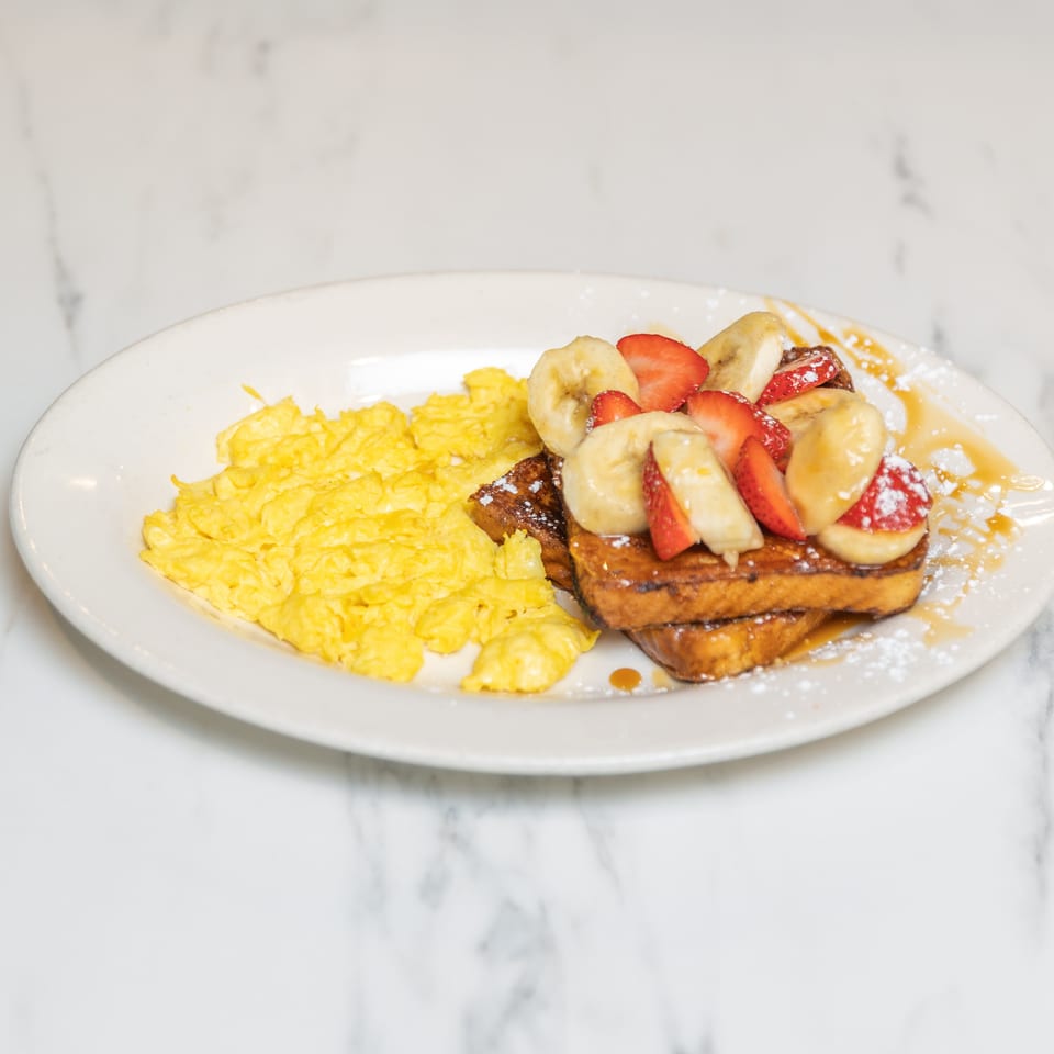 Scrambled eggs and French toast topped with strawberries, bananas, powdered sugar, and syrup.