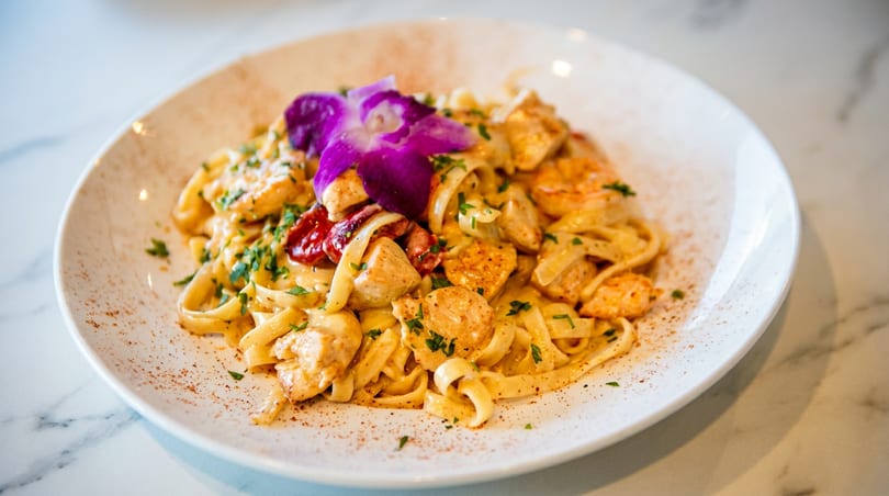 Creamy chicken fettuccine pasta with sun-dried tomatoes and a purple orchid garnish on a white plate.
