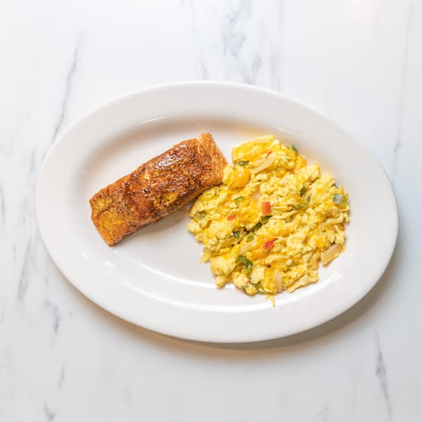 A white oval plate holds a seasoned salmon fillet and cheesy scrambled eggs with vegetables.