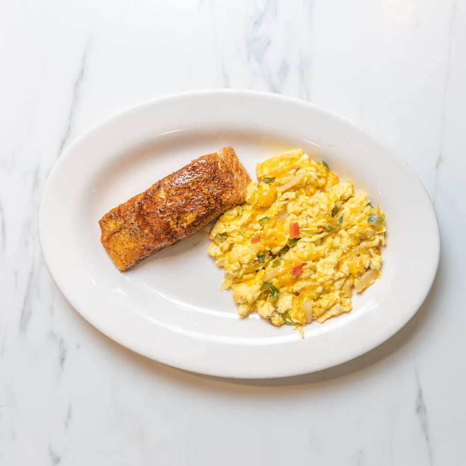 A white oval plate holds a seasoned salmon fillet and cheesy scrambled eggs with vegetables.