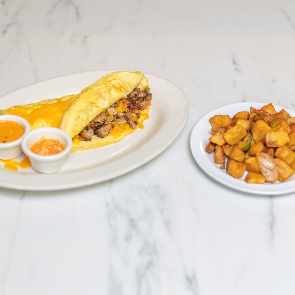 A steak and cheese omelet with two dipping sauces and a side of home fries on a marble surface.