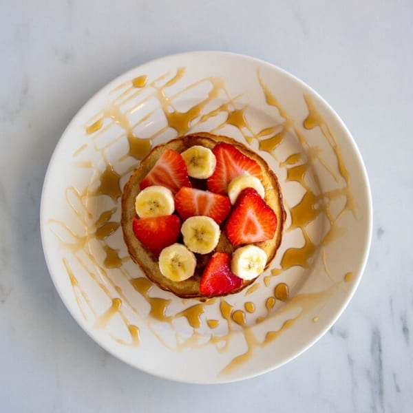 Single pancake topped with sliced strawberries and bananas, drizzled with syrup on a white plate.