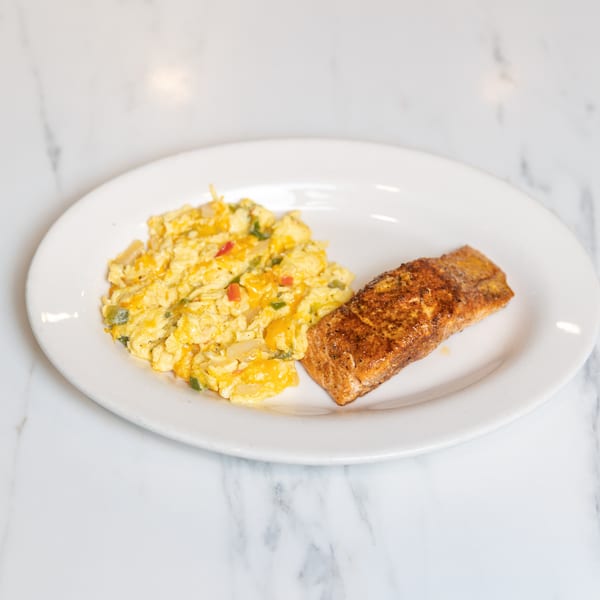 Scrambled eggs with cheese and vegetables next to a seasoned salmon fillet on a white plate.