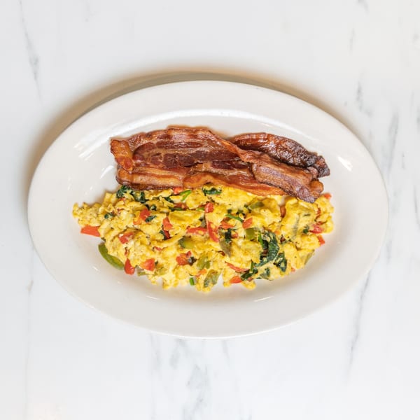 Scrambled eggs with colorful vegetables and two strips of crispy bacon on a white plate.