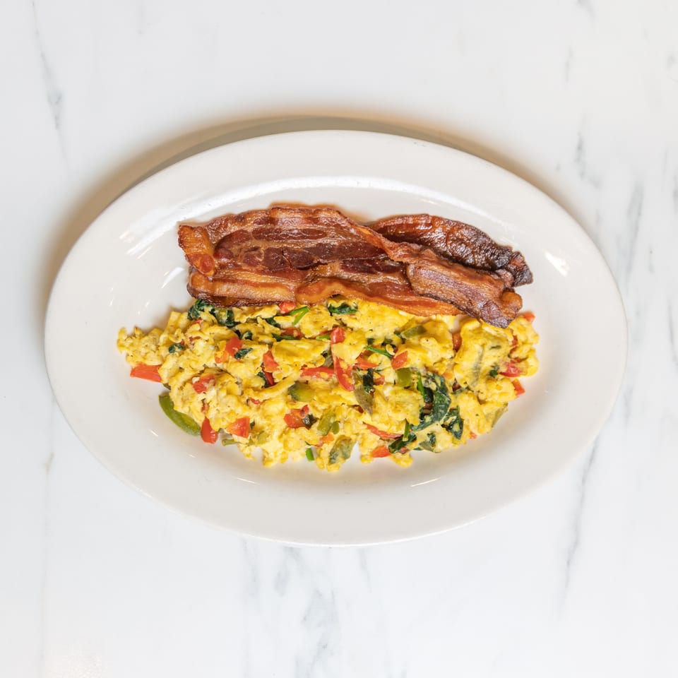 Scrambled eggs with colorful vegetables and two strips of crispy bacon on a white plate.