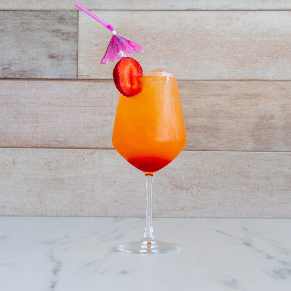 Vibrant orange and red cocktail with a strawberry, pink straw, and umbrella on marble.