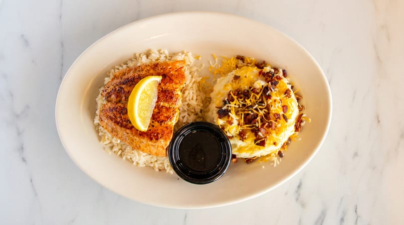 Grilled salmon with lemon, rice, mashed potatoes topped with cheese and bacon, and a side sauce.
