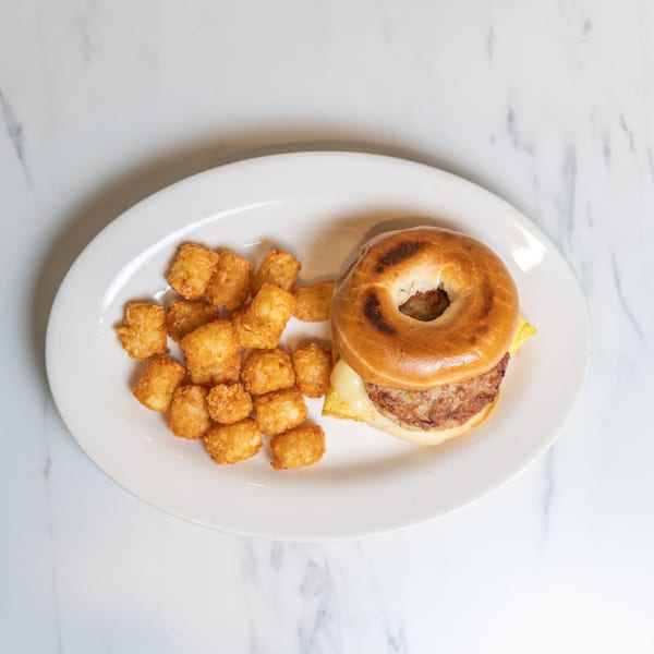 Breakfast bagel sandwich with sausage, egg, and cheese, served with a side of tater tots.