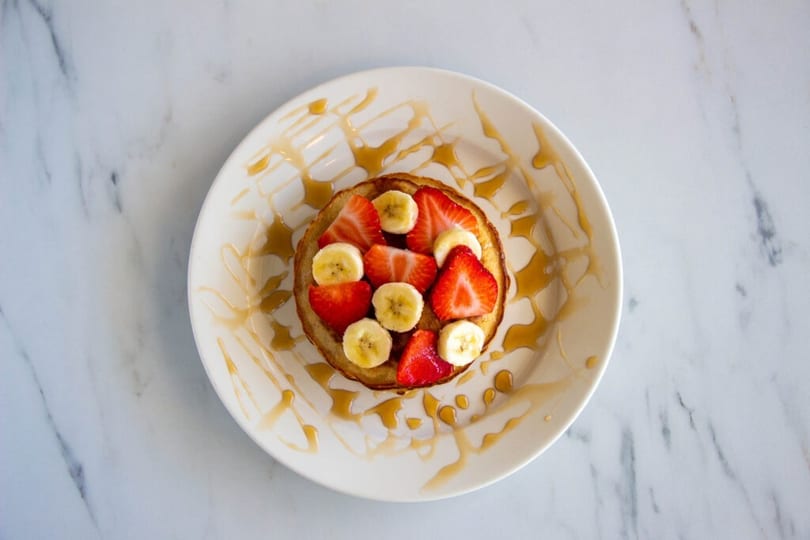 Single pancake topped with sliced strawberries and bananas, drizzled with syrup on a white plate.
