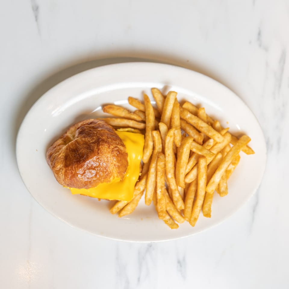 A golden-brown croissant sandwich with melted cheese and a side of crispy french fries on a white plate.